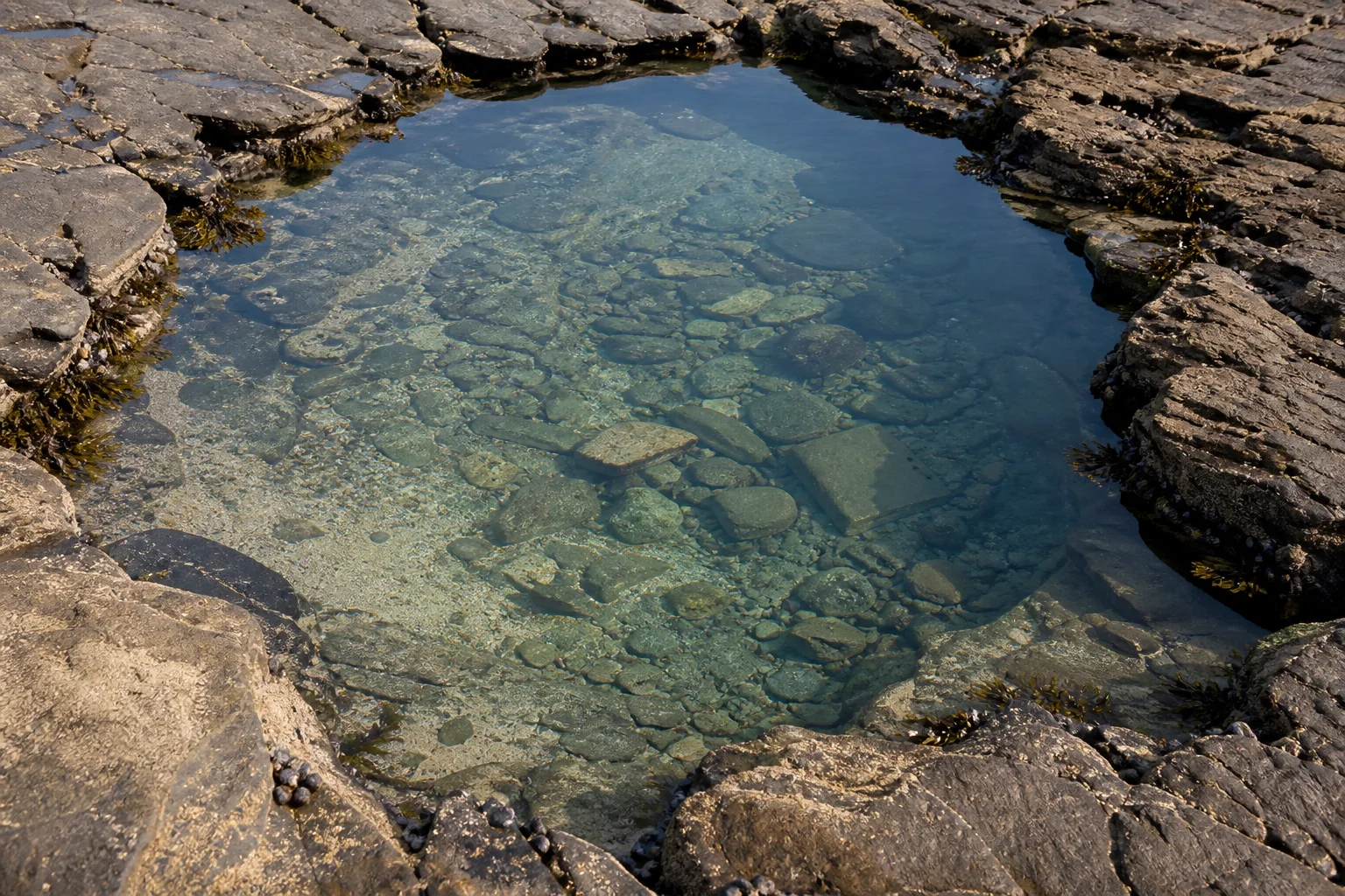 Clear seawater over a pebble bed, enclosed by eroded coastal rock shelves.
